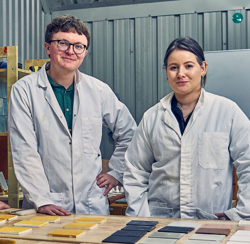CEO Dr Aled Roberts and a scientist, Raíssa Barroso, smiling in the lab, displaying a range of sustainable Eralith tiles on a bench in front of them.