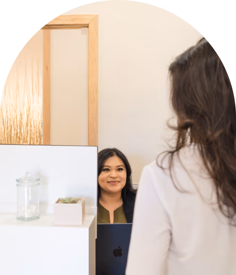 Daisy Vargas seated behind a white desk with a laptop, interacting with a standing visitor.