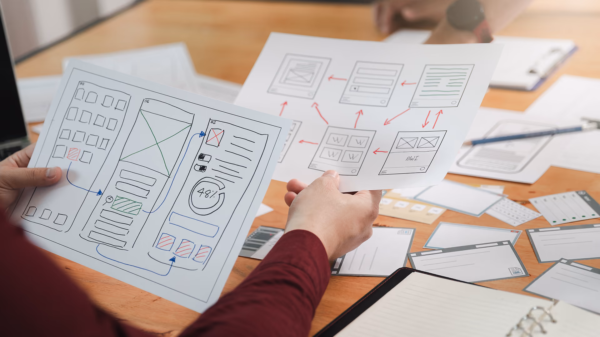 Ein Team begutachtet an einem Schreibtisch handgezeichnete App-Wireframes und Ablaufdiagramme; ausgebreitete Skizzen, Notizen und Bildschirmdarstellungen zeugen von kreativer Zusammenarbeit.