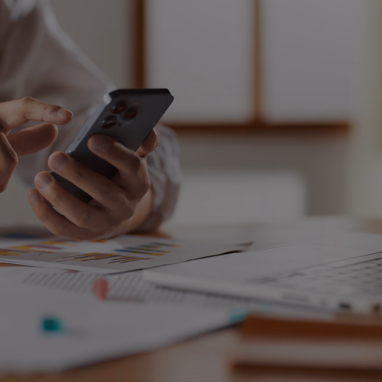 Eine Person nutzt an einem Schreibtisch ein Smartphone, umgeben von Diagrammen, Dokumenten und einem Stift; warmes natürliches Licht schafft eine konzentrierte, professionelle Atmosphäre.