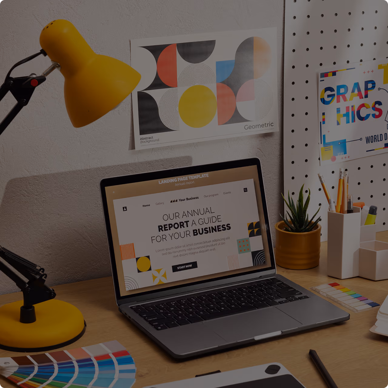 Creative home office desk with a yellow lamp, laptop showing an annual report template, geometric art, plant, stationery, and color swatches.
