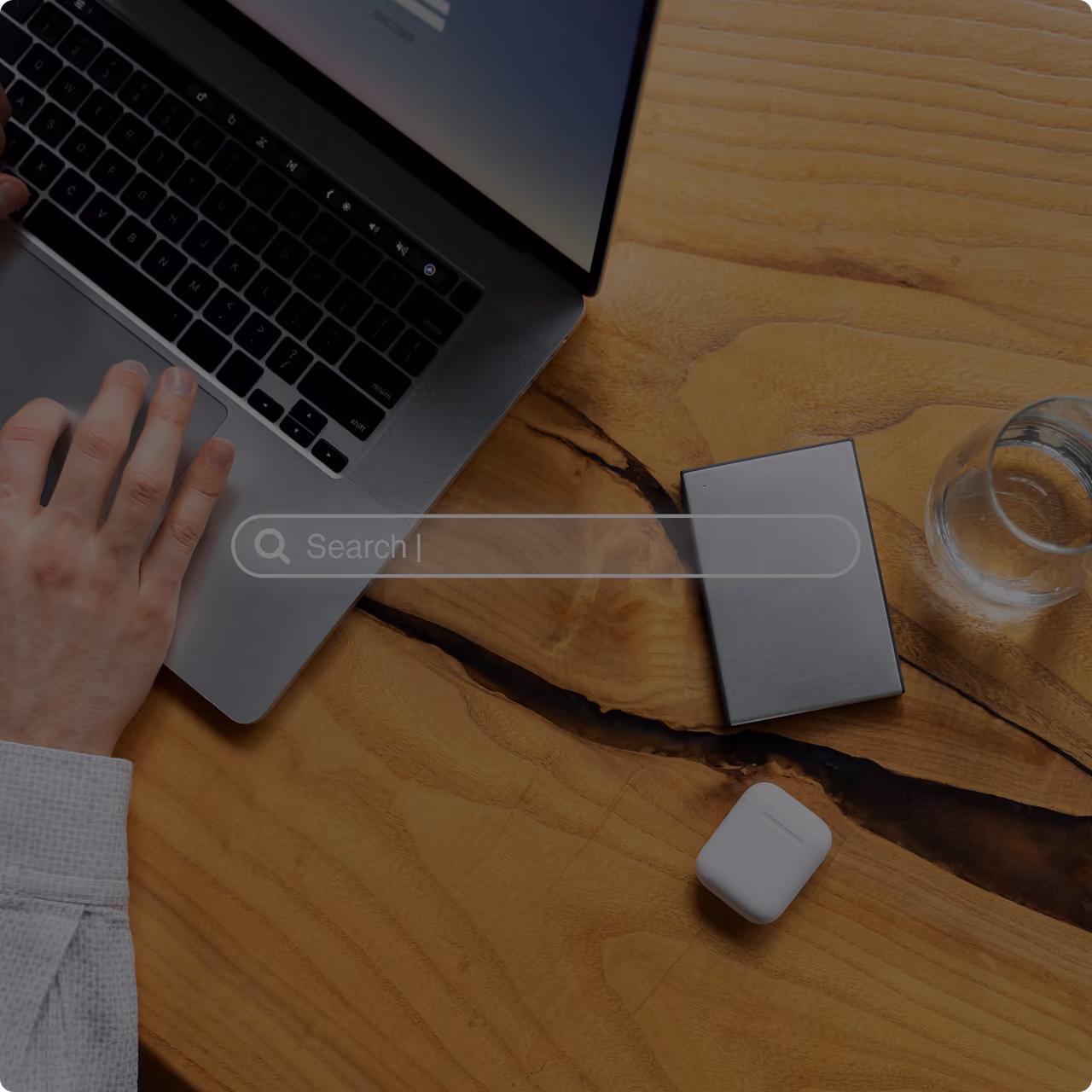 Ansicht von oben auf einen Arbeitsplatz mit Laptop, Hand auf der Tastatur, Smartphone, kabellosen Ohrhörern und Kaffee auf einem Holzschreibtisch; eine halbtransparente Suchleiste sorgt für eine moderne Note.