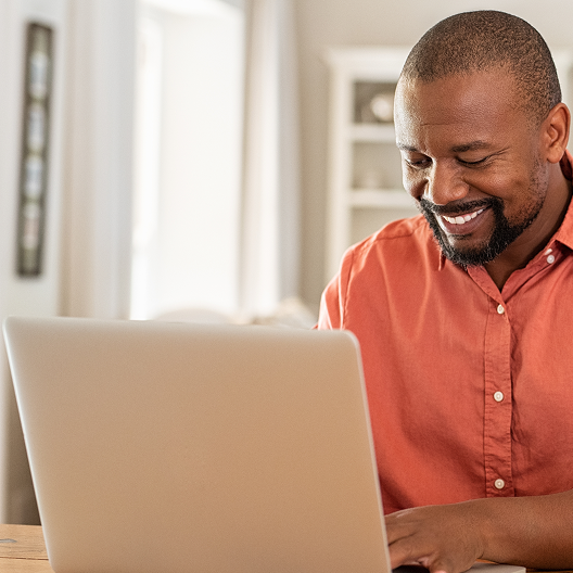 Smiling male church leader and pastor in an orange shirt working on a laptop at a wooden table indoors.