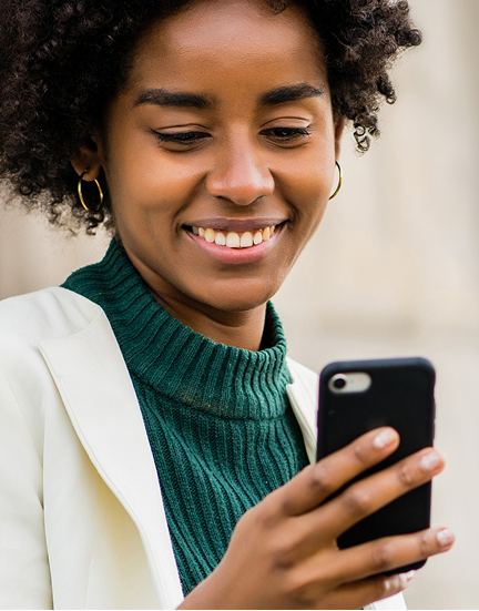 Smiling woman with curly hair wearing a green turtleneck and white jacket, looking at her mobile giving app on her phone.