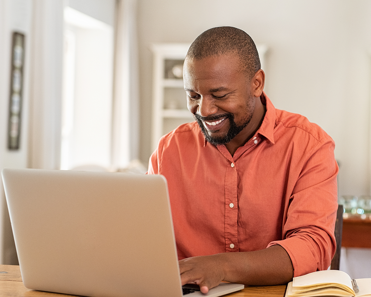 Smiling male church leader and pastor in an orange shirt working on a laptop at a wooden table indoors.