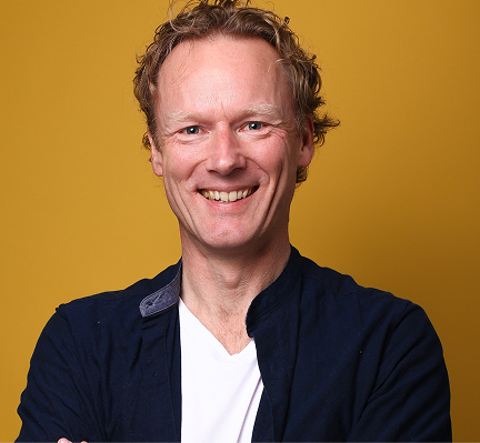 Smiling man with light brown curly hair wearing a white shirt and dark blue jacket on mustard background.