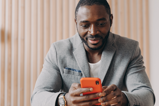 Smiling male church leader in gray blazer using an orange smartphone against a wood panel background.