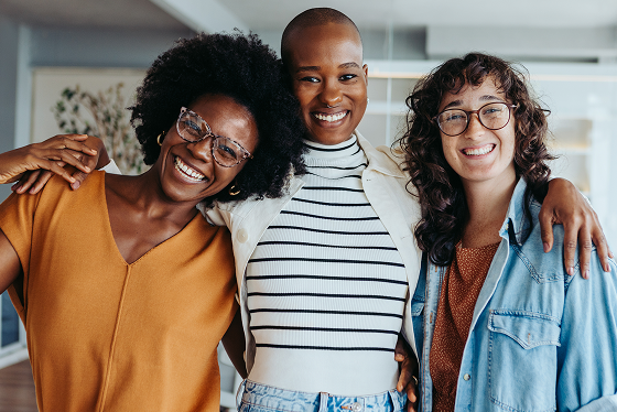 Three female faith-based church donors smiling with arms around each other in a bright indoor space.