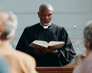 Male church leader and pastor in black robe reading from an open Bible during a church service.