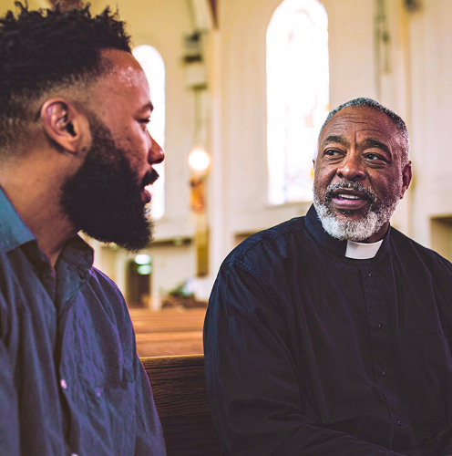 A male church leader and pastor and a congregant sitting in a church pew having a conversation.
