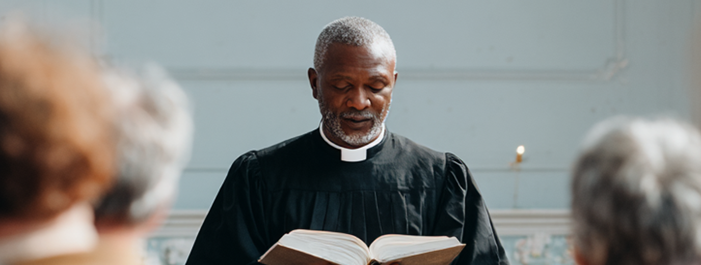 Male church leader and pastor in black robe reading from an open Bible during a church service.