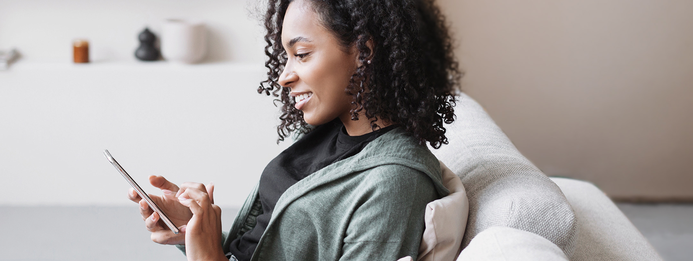 Smiling female church donor with curly hair using a digital giving tool on her mobile phone while seated indoors on a couch.