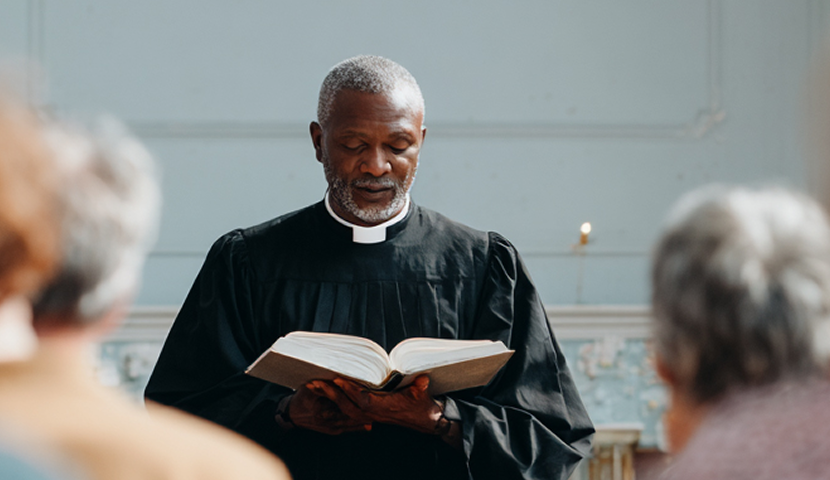 Male church leader and pastor in black robe reading from an open Bible during a church service.