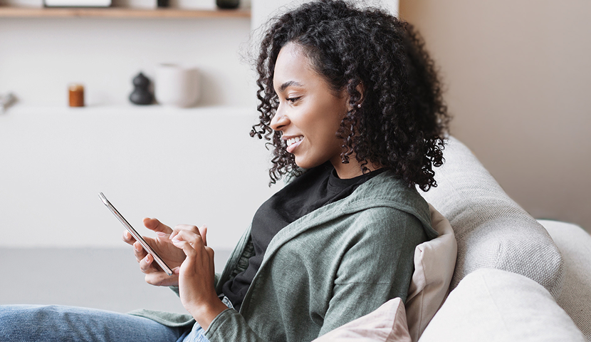 Smiling female church donor with curly hair using a digital giving tool on her mobile phone while seated indoors on a couch.