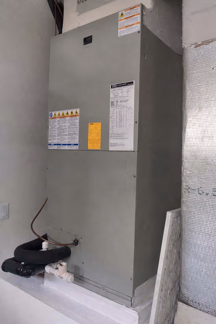 Gray HVAC unit installed against a wall with attached pipes and warning labels.