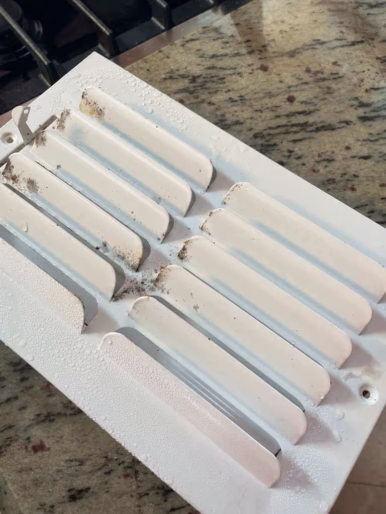 White metal air vent cover with condensation droplets and visible dirt on its slats, placed on a granite countertop.