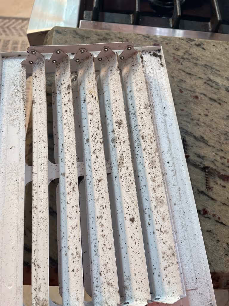Close-up of a white metal air vent grille with dust and dirt accumulation on its slats placed on a kitchen countertop.