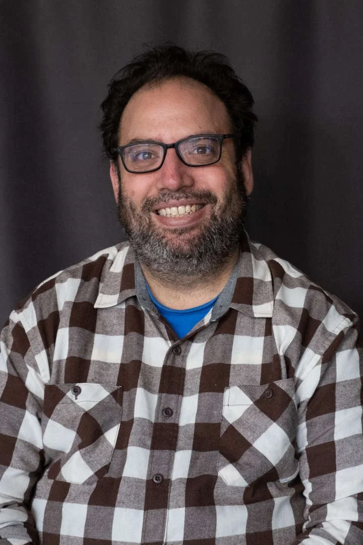 Staff photo of B Goldman smiling in plaid shirt