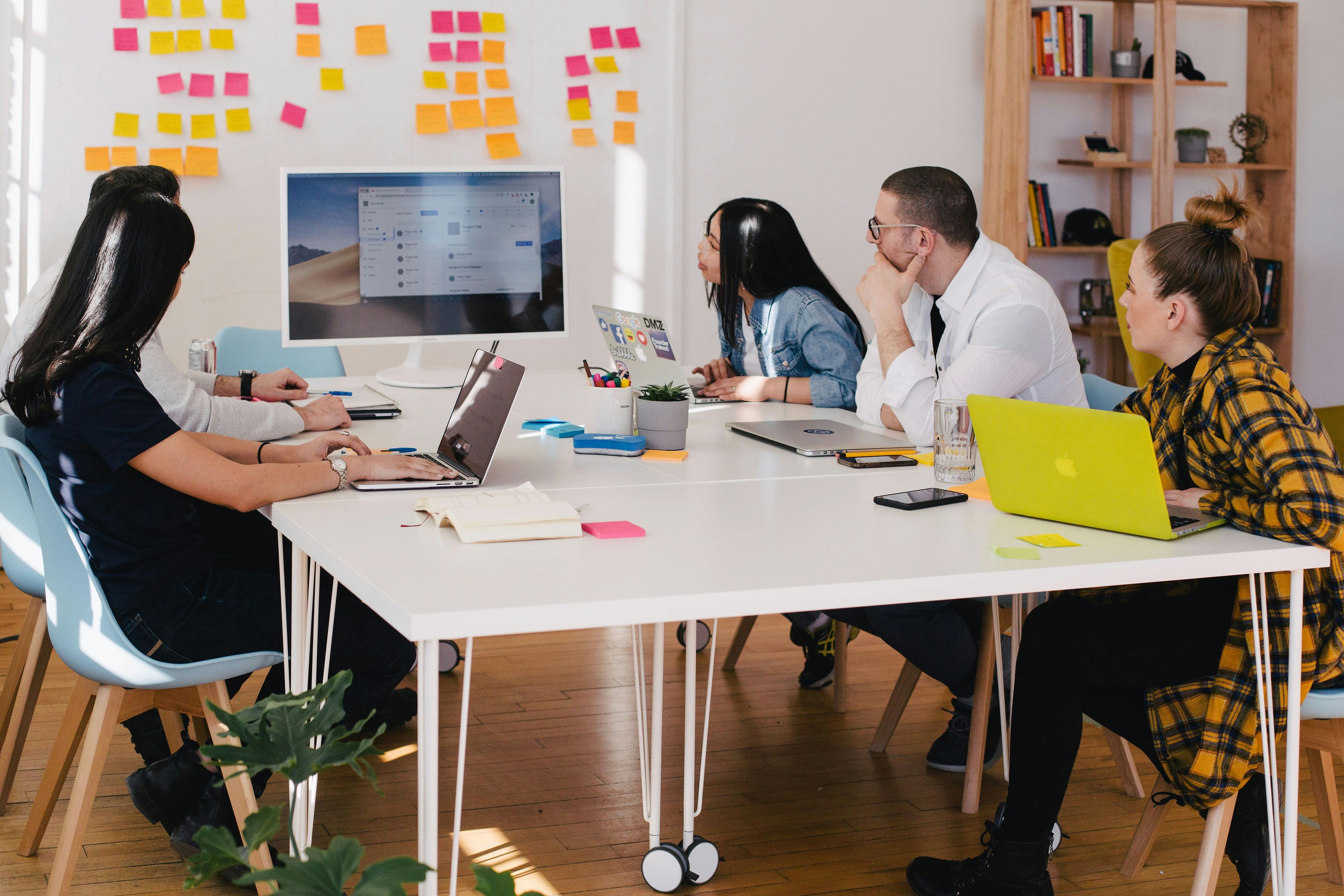 Five people seated around a table looking at a computer monitor with sticky notes on the wall behind them in a bright office.