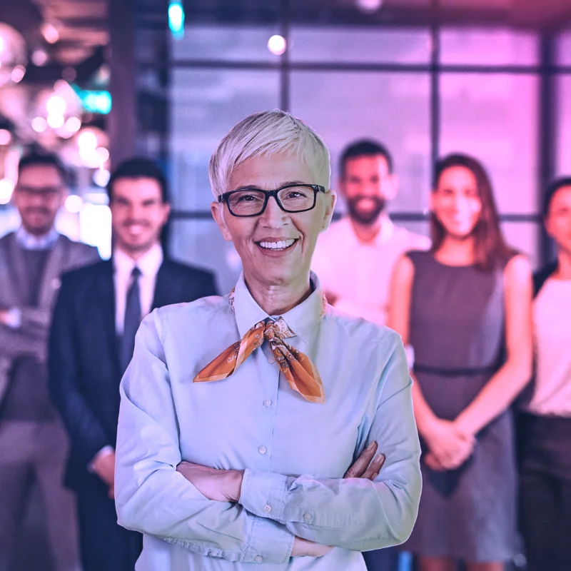 Führungskraft steht lächelnd im Vordergrund eines Teams in einem modernen Büro mit violettem Licht.