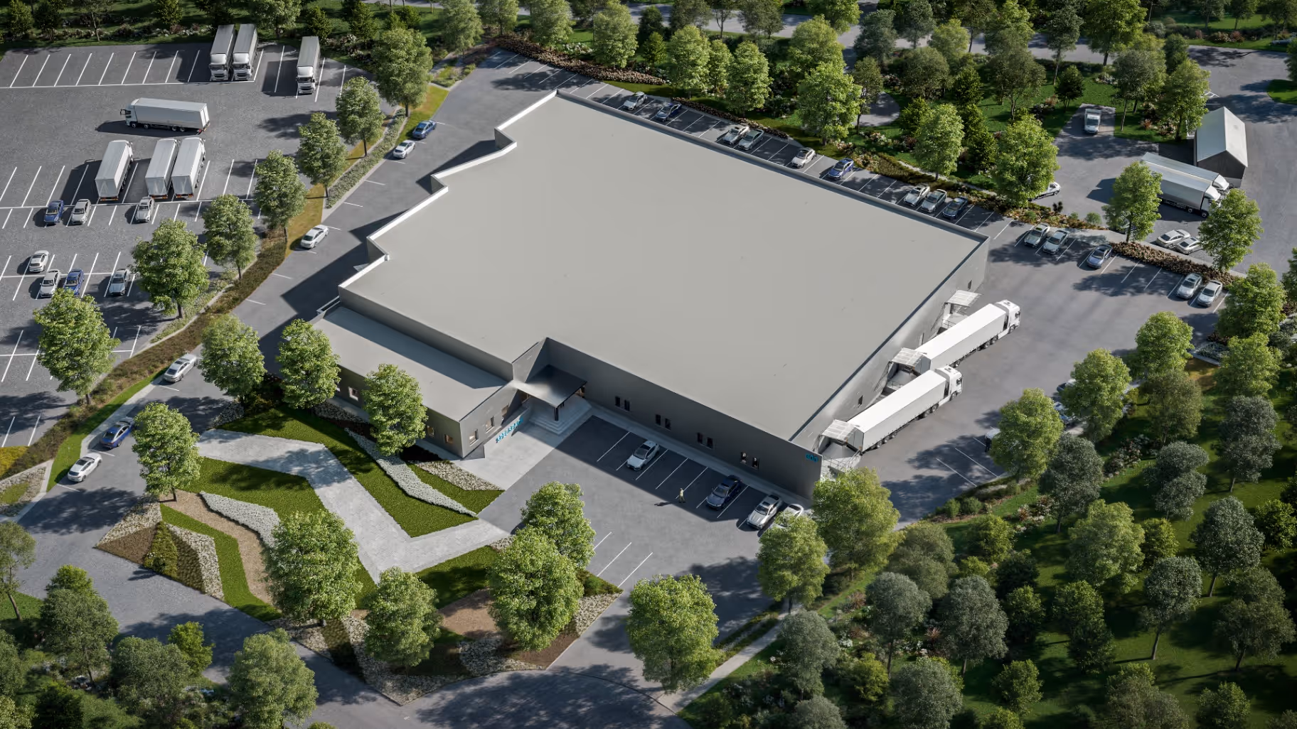 Aerial view of a large industrial warehouse surrounded by trees, with multiple trucks parked at loading docks and cars in parking lots.