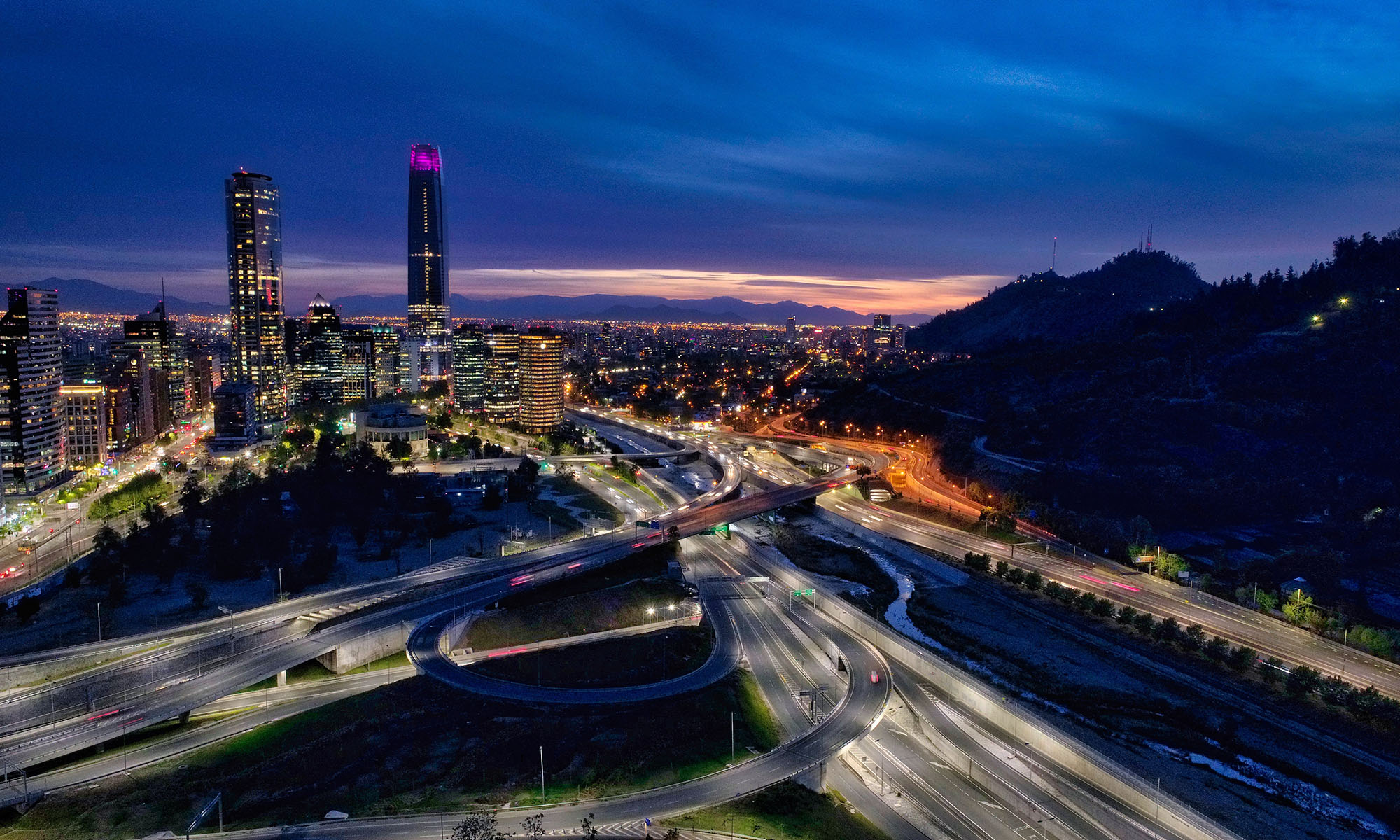 Nighttime cityscape with illuminated skyscrapers, highways with moving car lights, and mountains in the background under a cloudy sky.