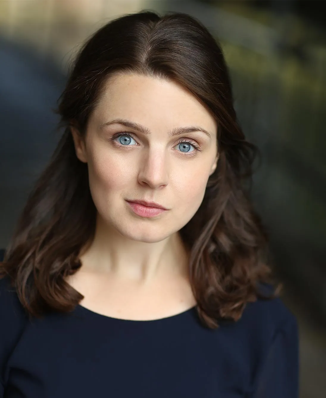 Close-up portrait of a woman with blue eyes and brown hair wearing a dark top against a softly blurred background.