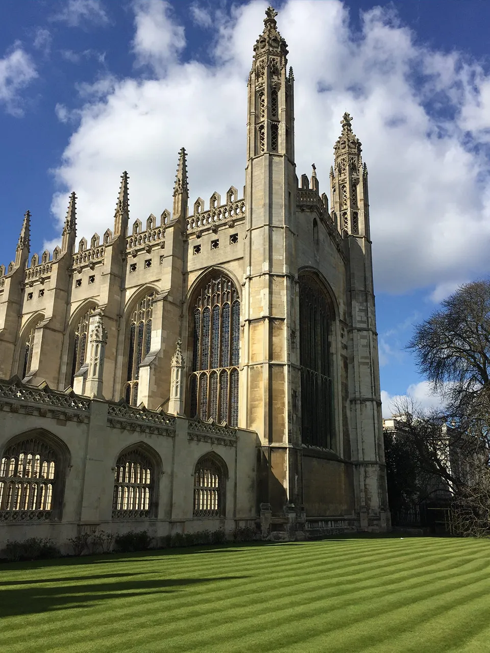 Gothic-style stone building with tall ornate windows and spires, beside a neatly striped green lawn under a partly cloudy sky.