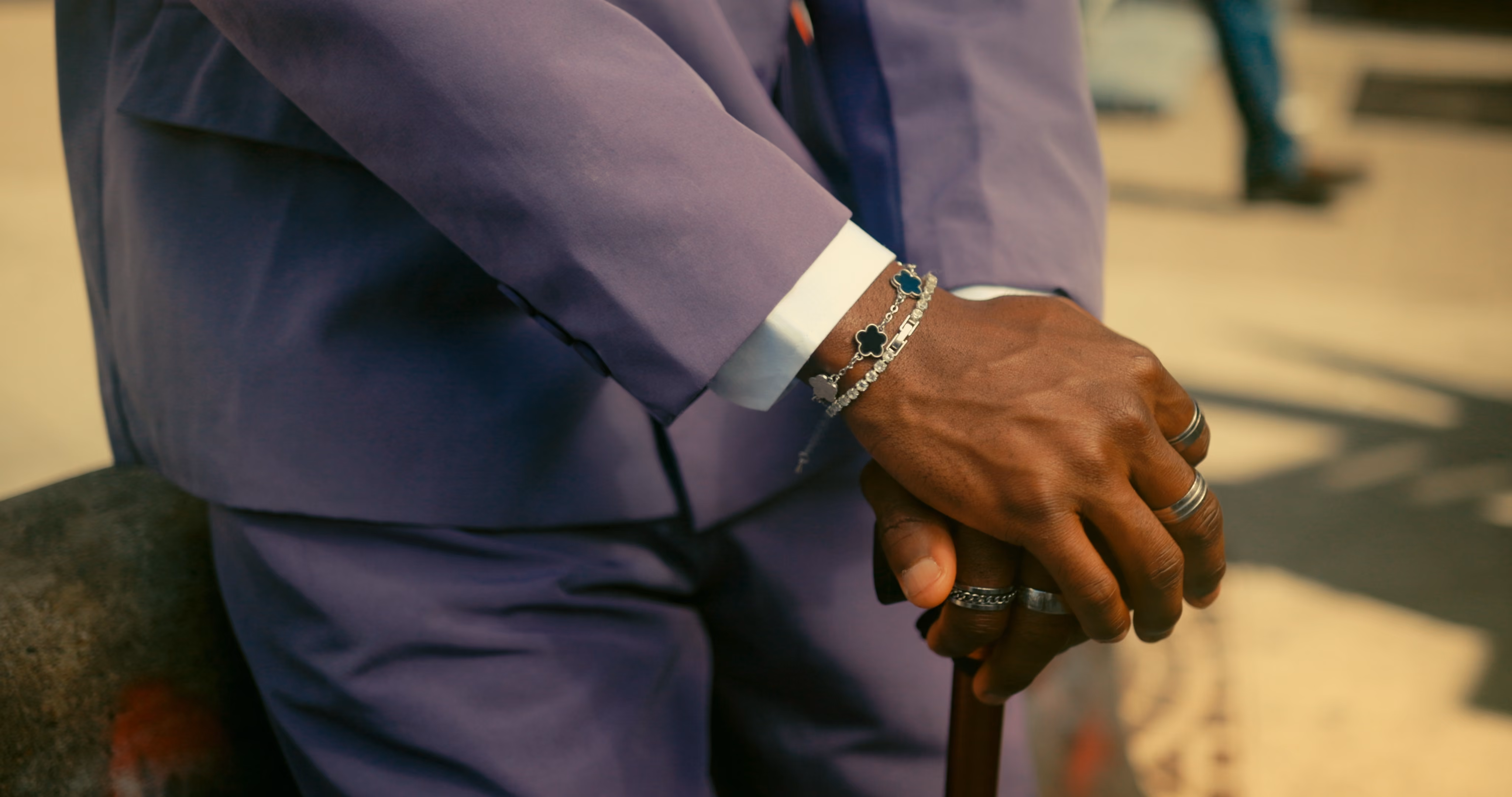 Close-up of hands with rings and bracelets holding a cane, dressed in a purple suit.