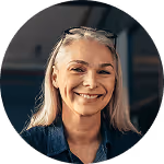 Ältere Frau mit grauem Haar und Brille lächelnd, trägt blaues Hemd in Innenraum.
