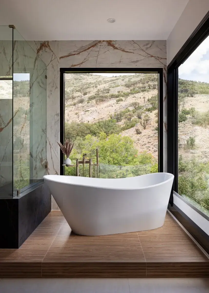 Modern white freestanding tub on wood platform with large windows showing hillside greenery.