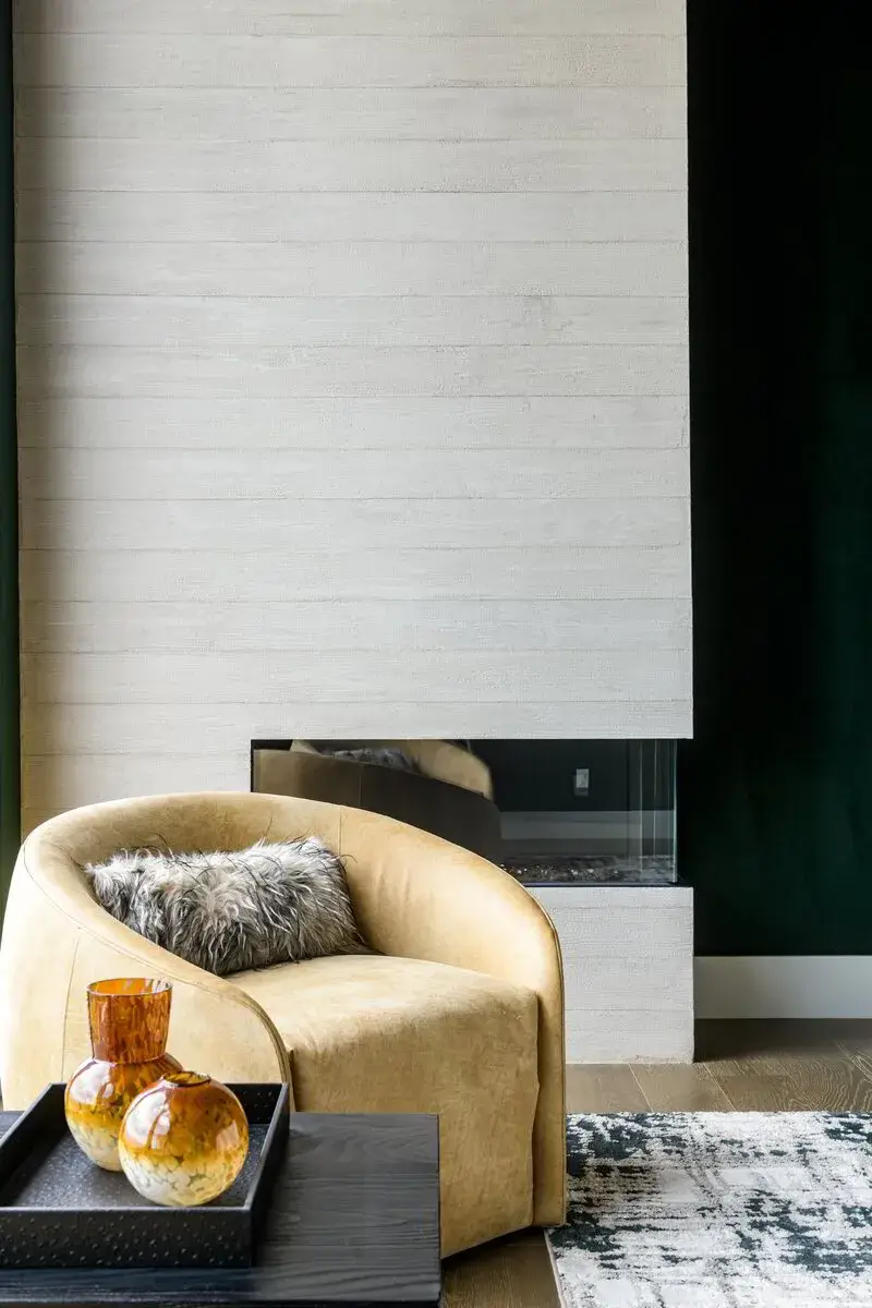 Modern beige armchair with a fluffy pillow near a white fireplace and dark wall in a living room.