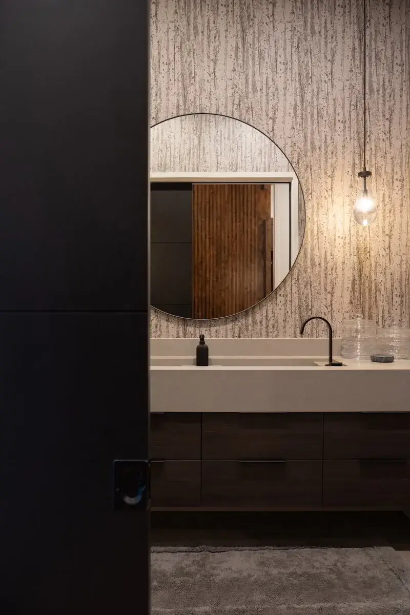 Modern bathroom vanity with round mirror, black faucet, pendant light, and textured wallpaper.