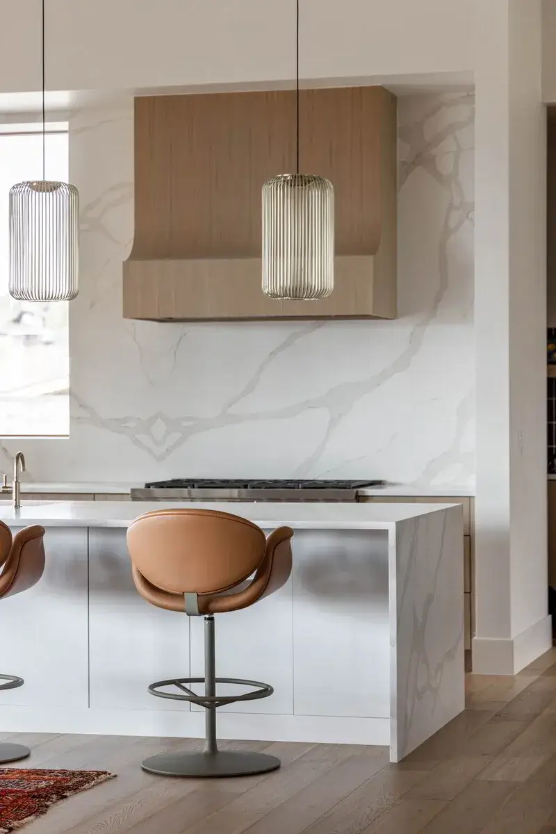 Modern kitchen with marble island, leather bar stools, wood range hood, and hanging pendant lights.
