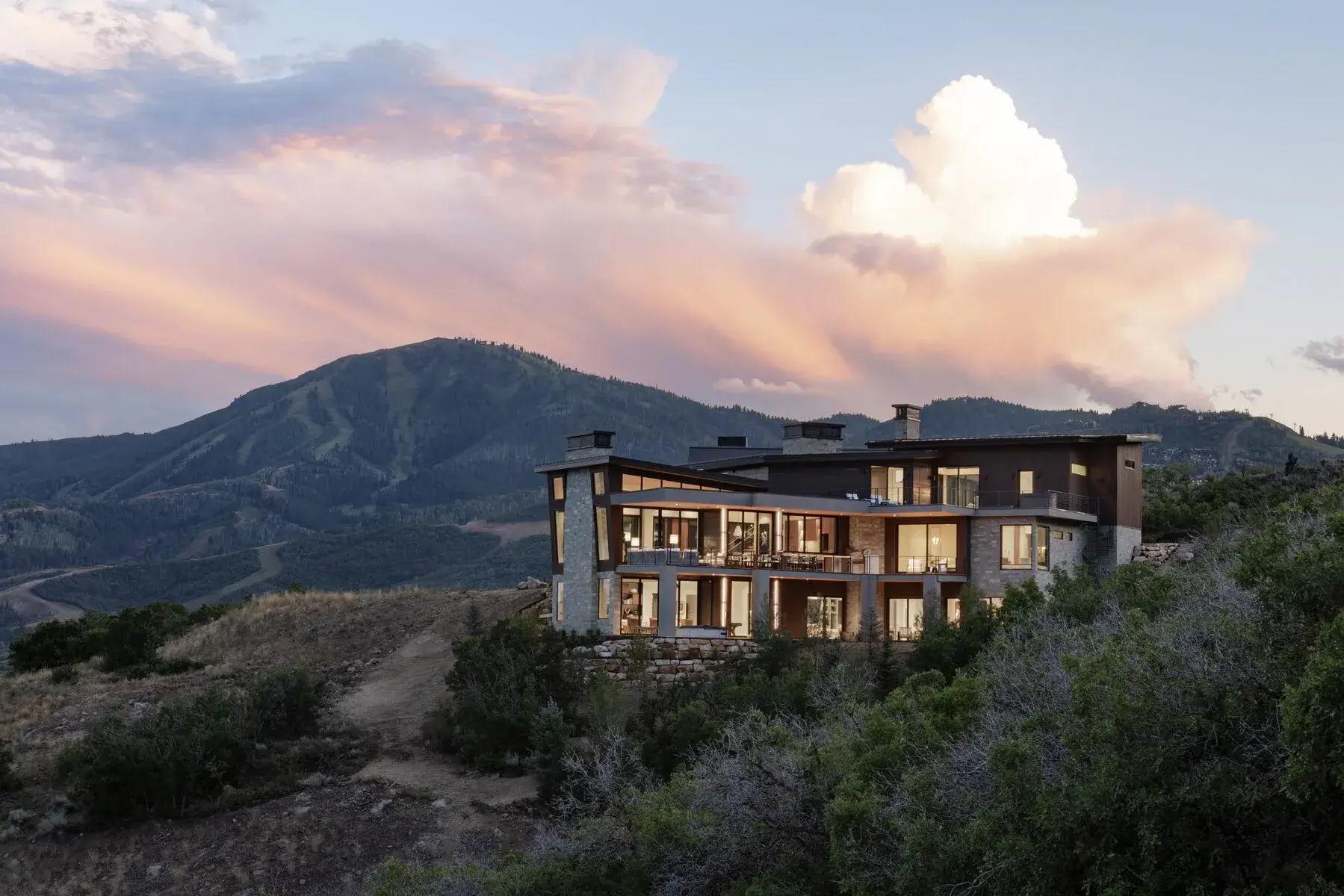 Modern house with large windows lit inside on hillside at sunset with mountain and colorful sky.
