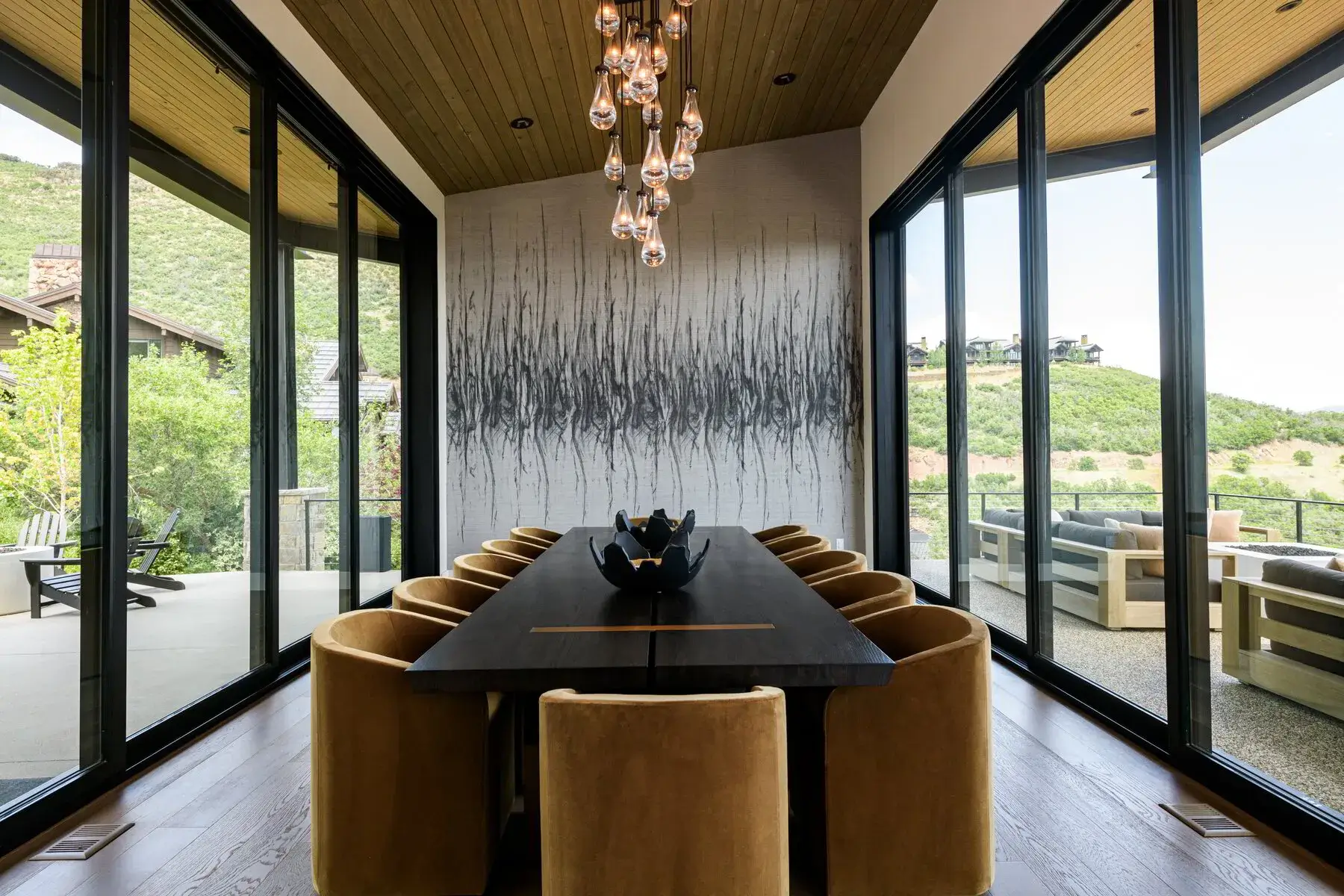 Modern dining room with large dark table, mustard chairs, pendant lights, and mountain views through glass walls.