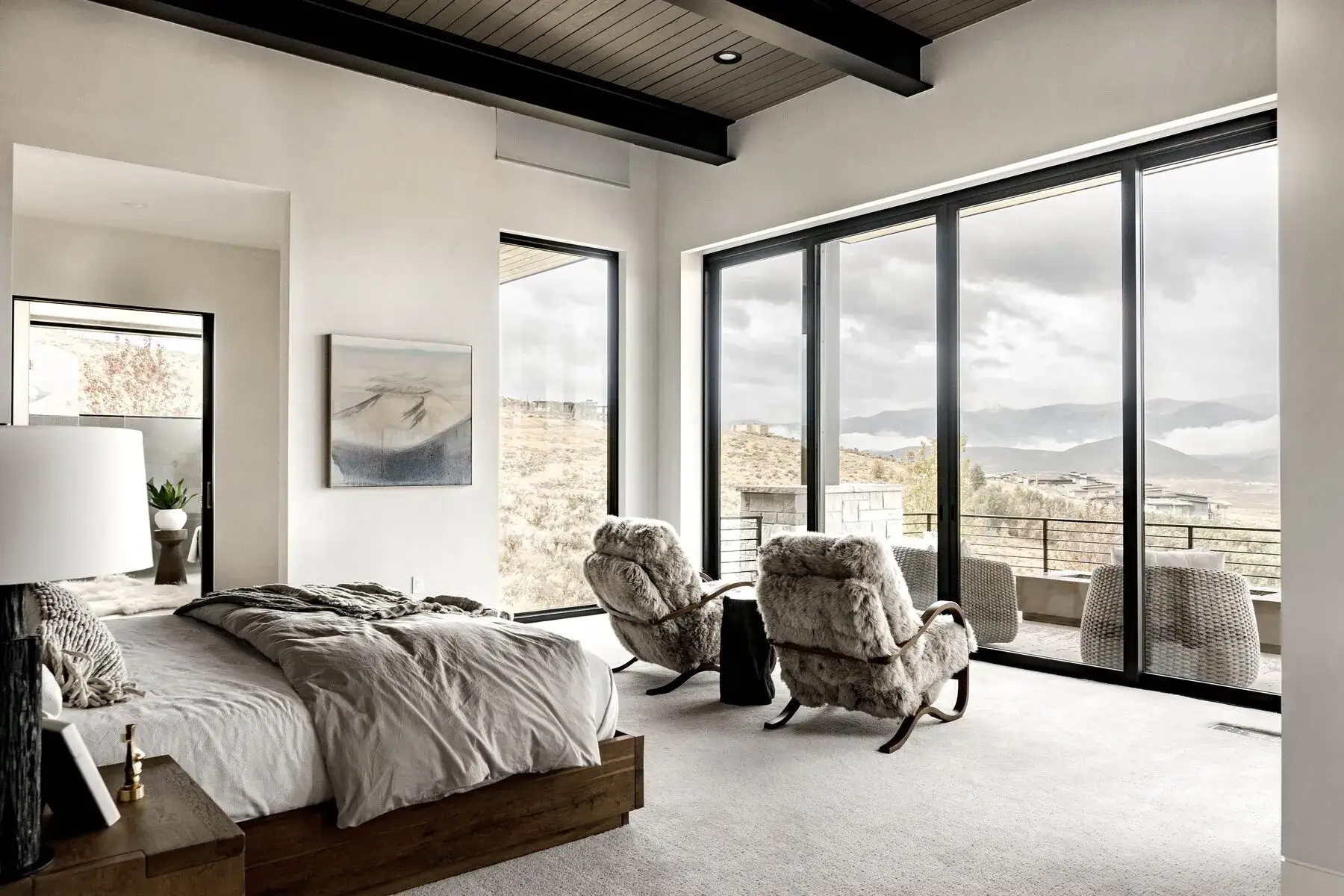Modern bedroom with a wooden bed, furry chairs, and large windows overlooking a mountainous view.