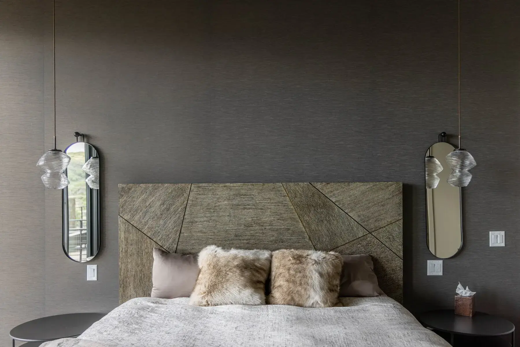 Modern bedroom with textured headboard, two fur pillows, soft lamps, and oval mirrors on dark walls.