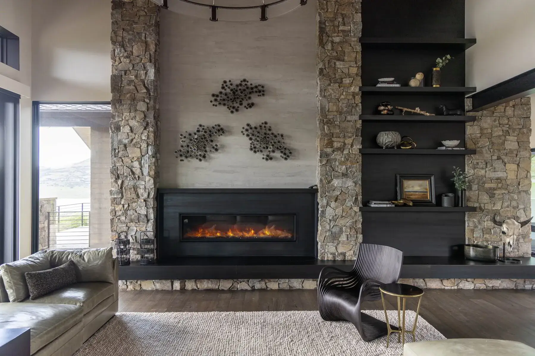 Modern living room with stone fireplace, black shelves, beige sofa, black chair, and a small round table.