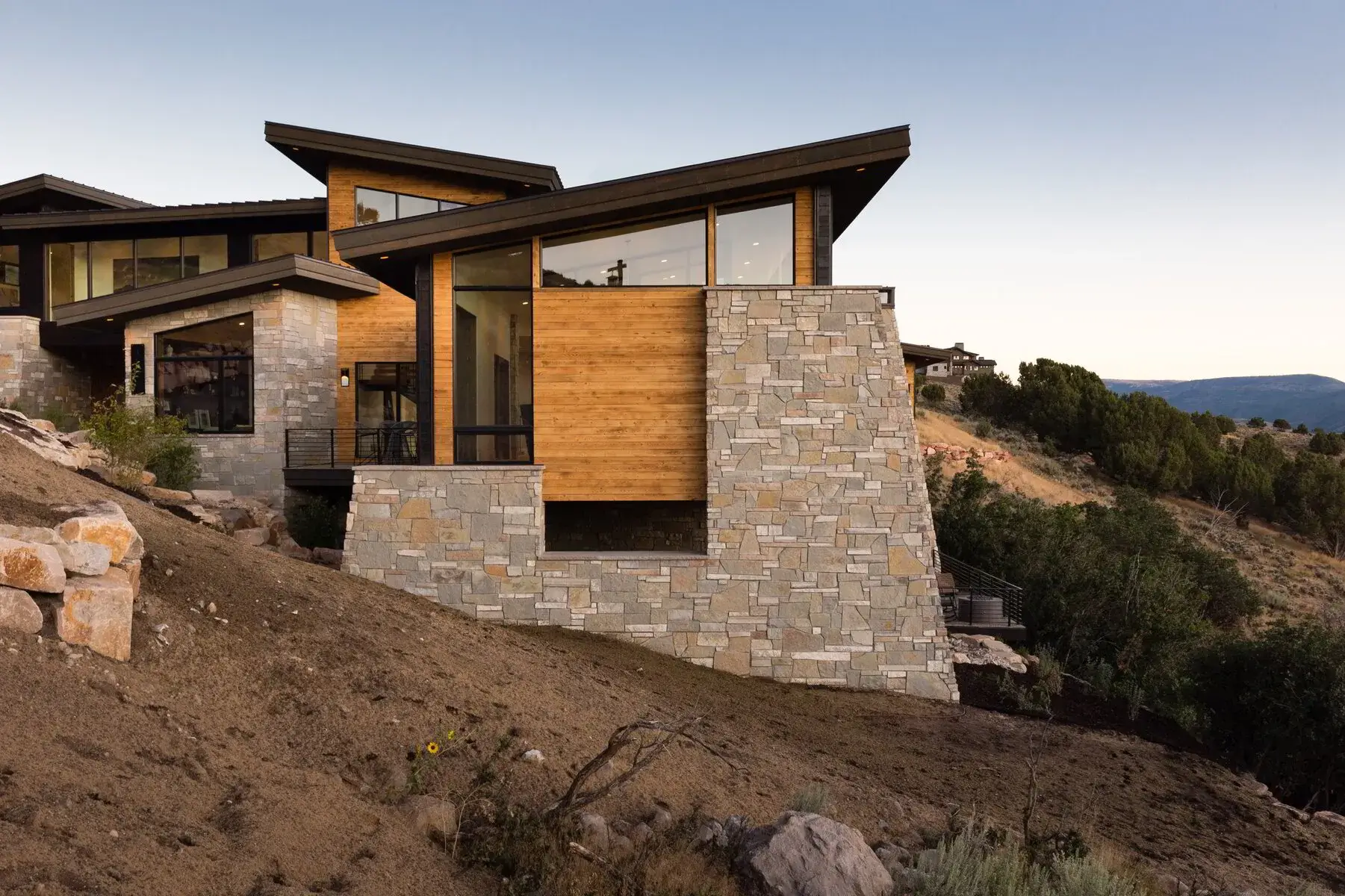 Modern house with stone and wooden exterior on a hillside with trees and mountains in the background.