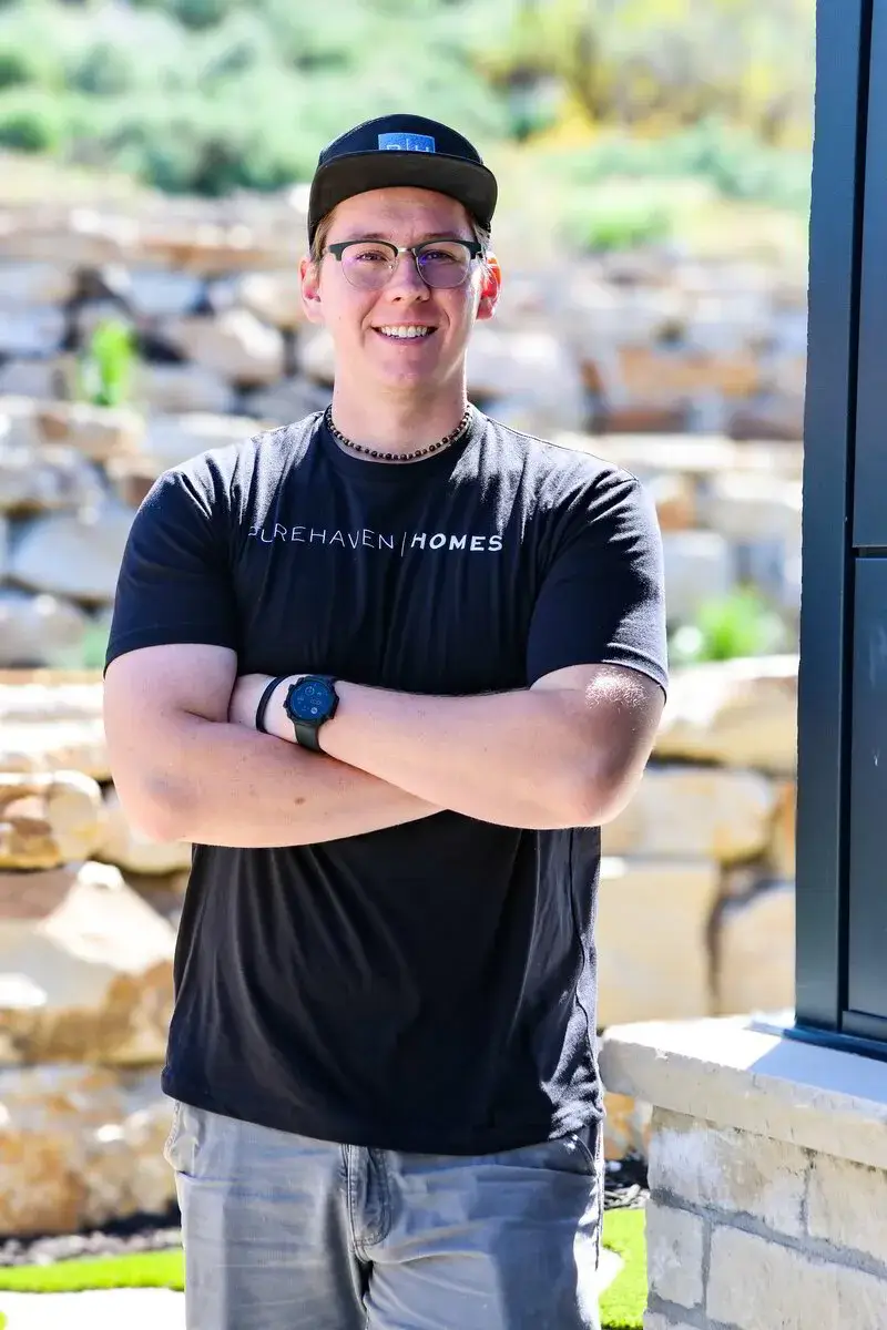 Smiling man in glasses and cap wearing a black Purehaven Homes shirt with arms crossed outdoors.
