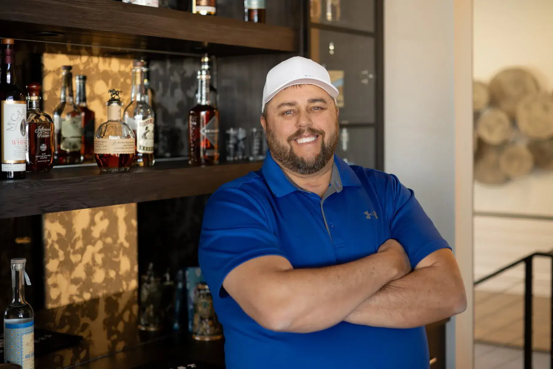 Smiling man in blue polo and white cap standing with arms crossed by a shelf of liquor bottles.
