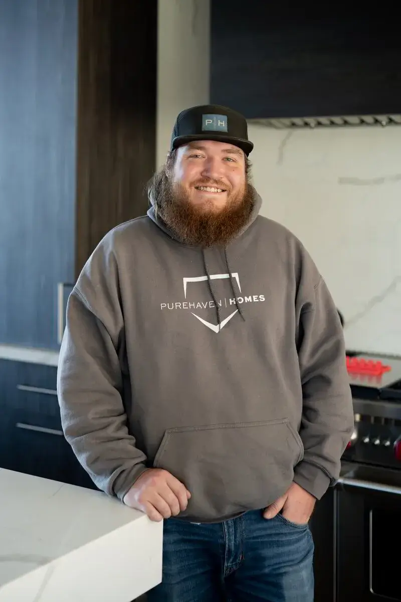 Smiling bearded man wearing a grey Purehaven Homes hoodie and cap, standing in a modern kitchen.