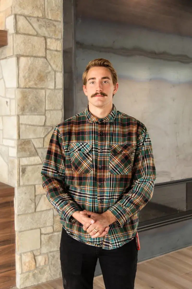 Man with a mustache wearing a brown and green plaid shirt standing indoors with stone wall.