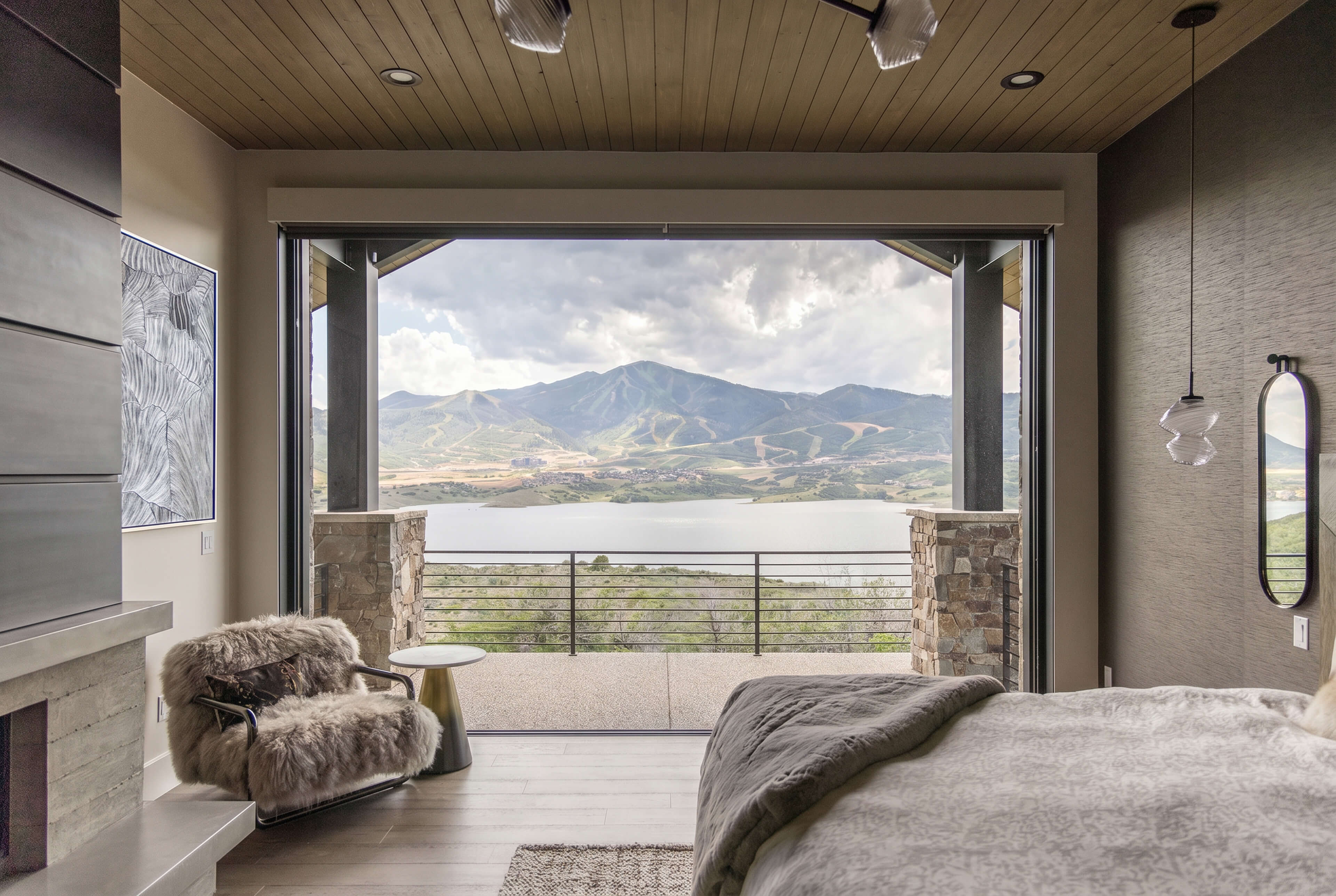 Modern bedroom with fur chair and large open window showing lake and mountain views.