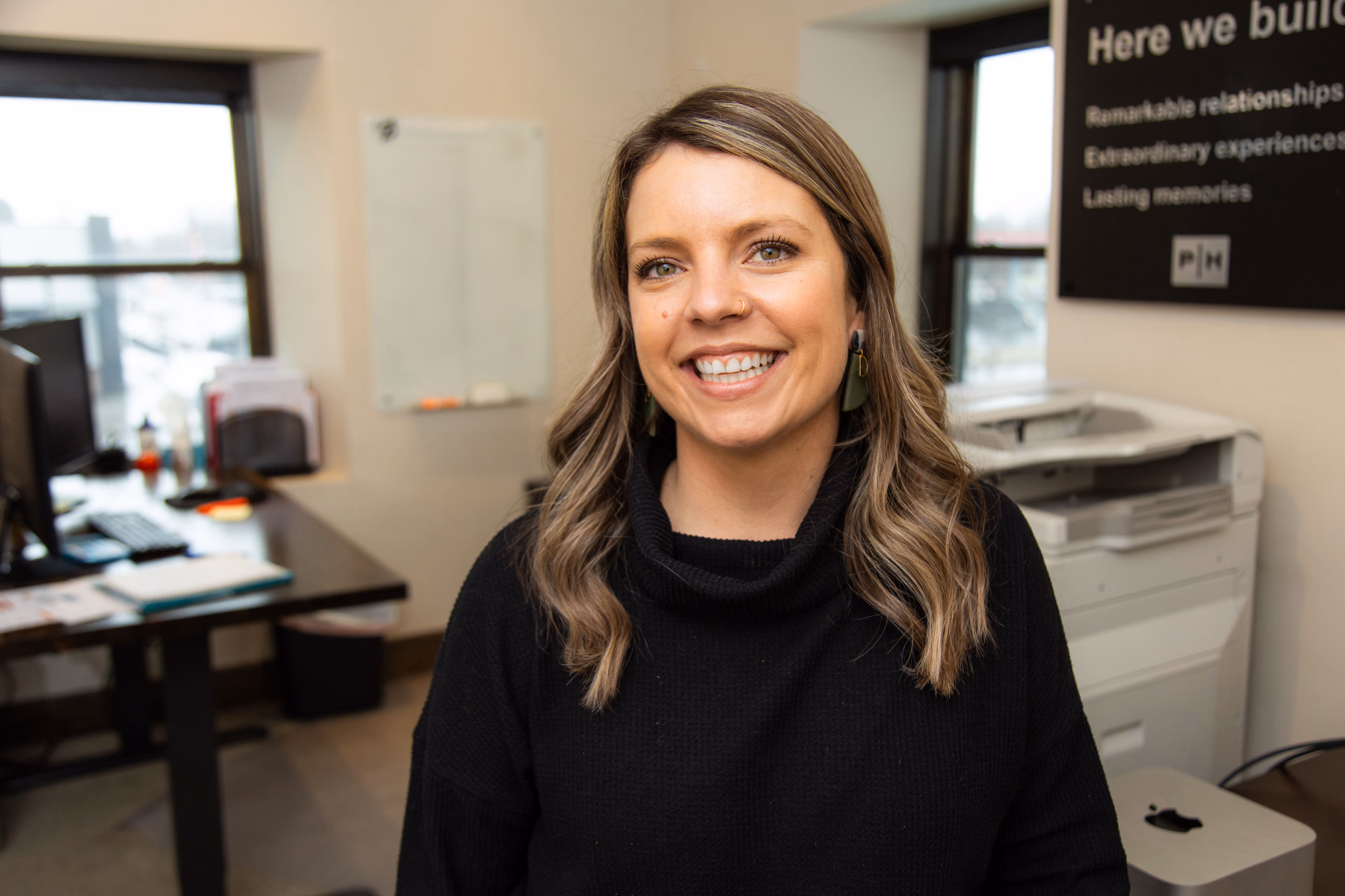 Smiling woman with shoulder-length hair in black sweater in an office with desks and a printer.