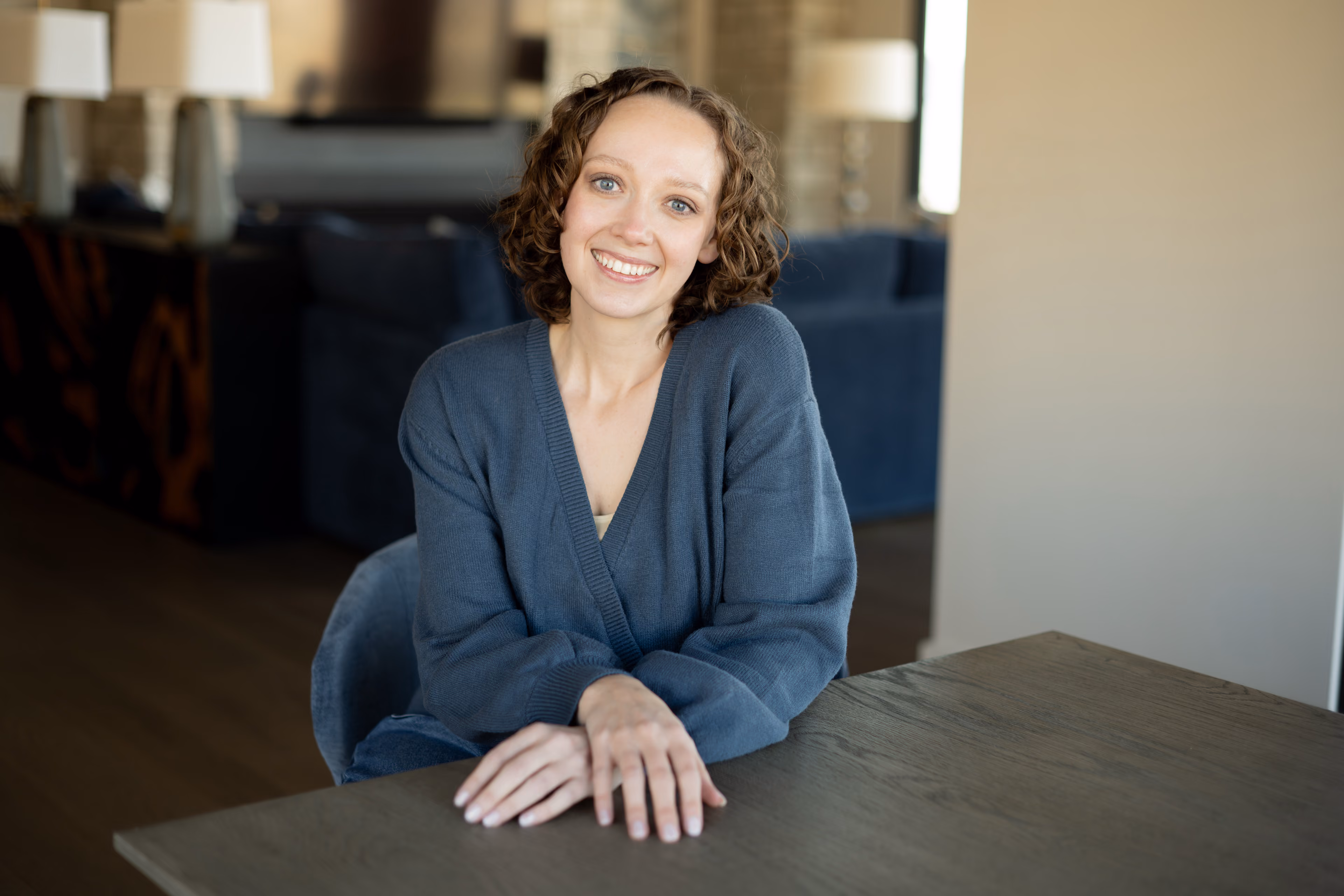 Smiling woman with curly hair wearing a blue sweater sitting at a wooden table indoors.