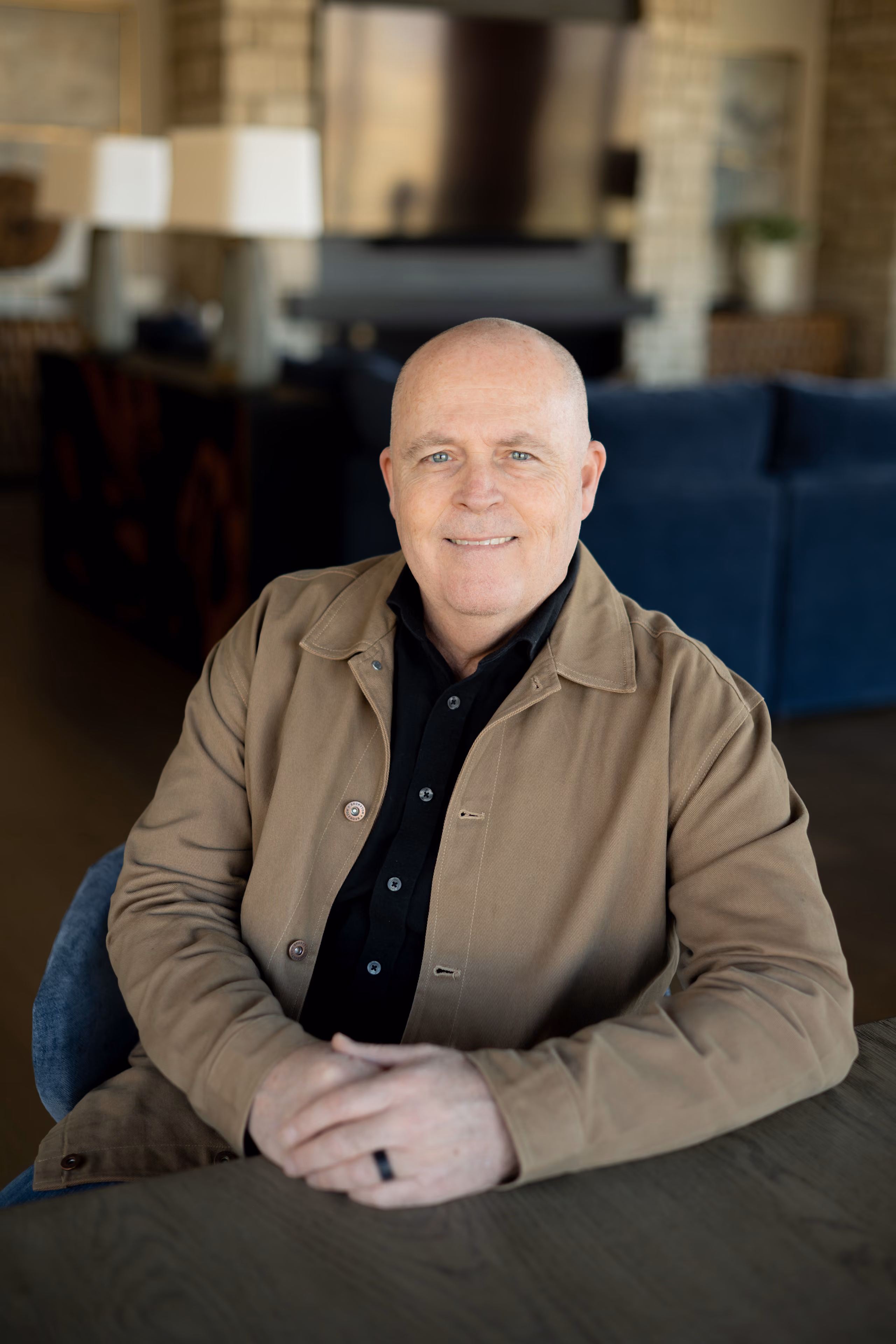 Bald man in tan jacket and black shirt sitting at table with hands clasped, smiling at camera indoors.