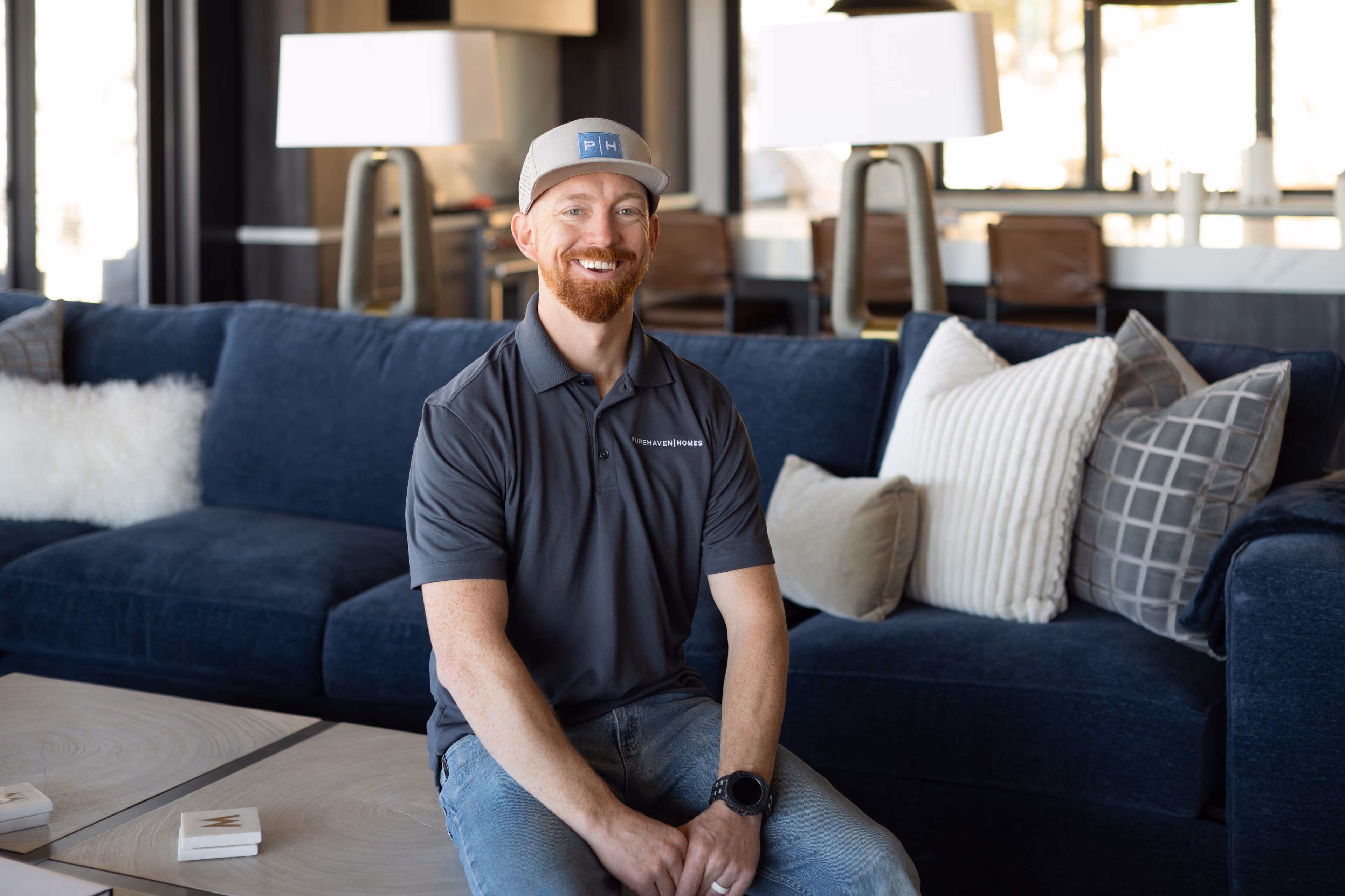 Smiling man with beard and cap sitting on a blue couch with gray and white pillows in bright room.