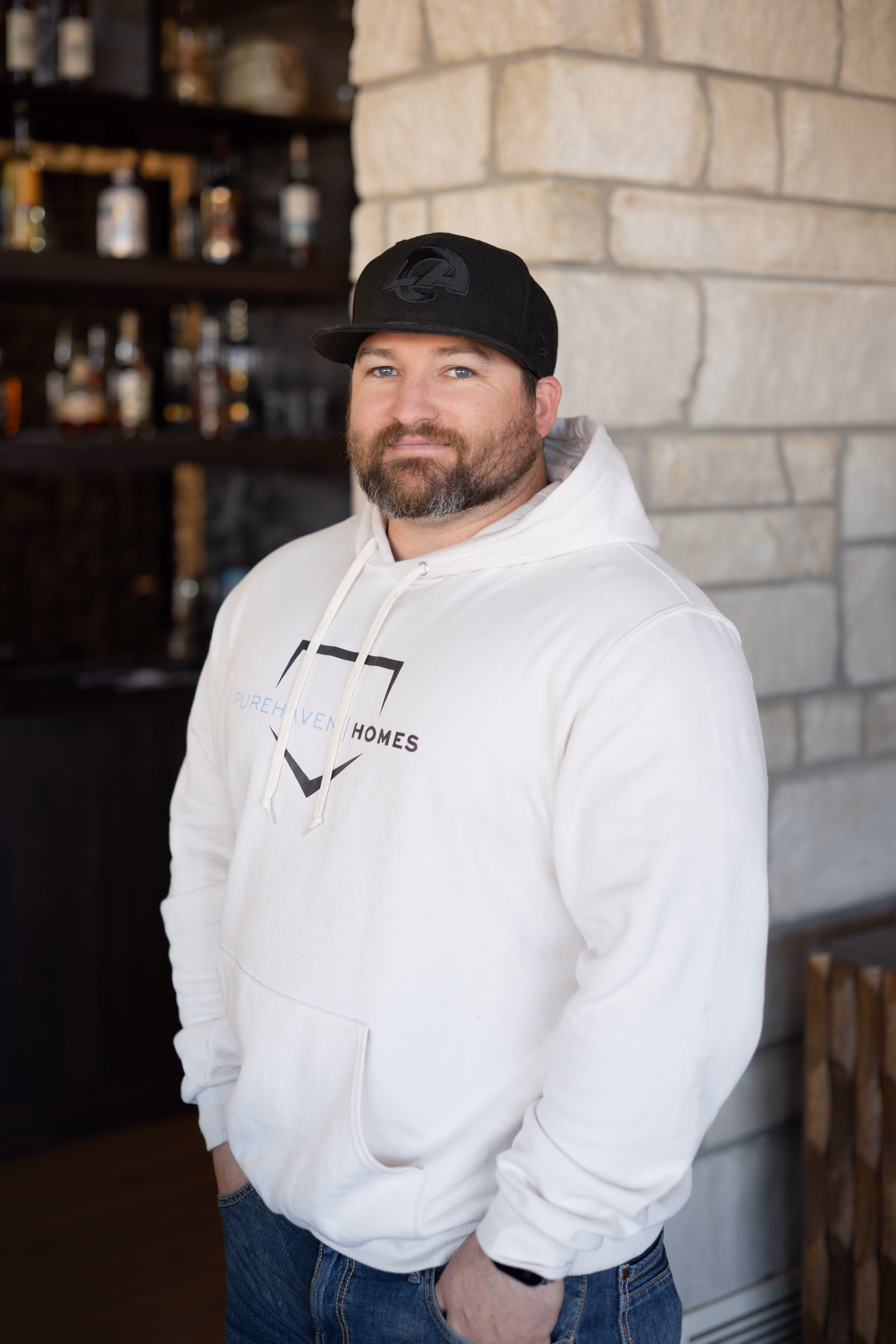 Man with beard wearing white Pure Haven Homes hoodie and black cap standing indoors by a stone wall.